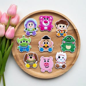 May include: A collection of embroidered patches featuring characters from the animated film Toy Story. The patches include Buzz Lightyear, Lotso, Woody, and others, all with detailed stitching and vibrant colors. They are arranged on a wooden tray.