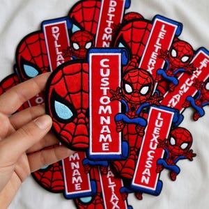 May include: A collection of red, blue, and white Spider-Man patches. Some patches feature the Spider-Man mask, while others display the character in full. Several patches have the text "CUSTOM NAME" or other names.