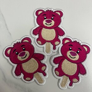 K&ouml;nnte beinhalten: Drei gestickte Aufn&auml;her mit einem rosa Teddyb&auml;r-Design mit beigem Bauch und einem Eisstiel. Der B&auml;r hat ein l&auml;chelndes Gesicht und einen wei&szlig;en Umriss. Die Aufn&auml;her befinden sich auf einer wei&szlig;en Oberfl&auml;che.