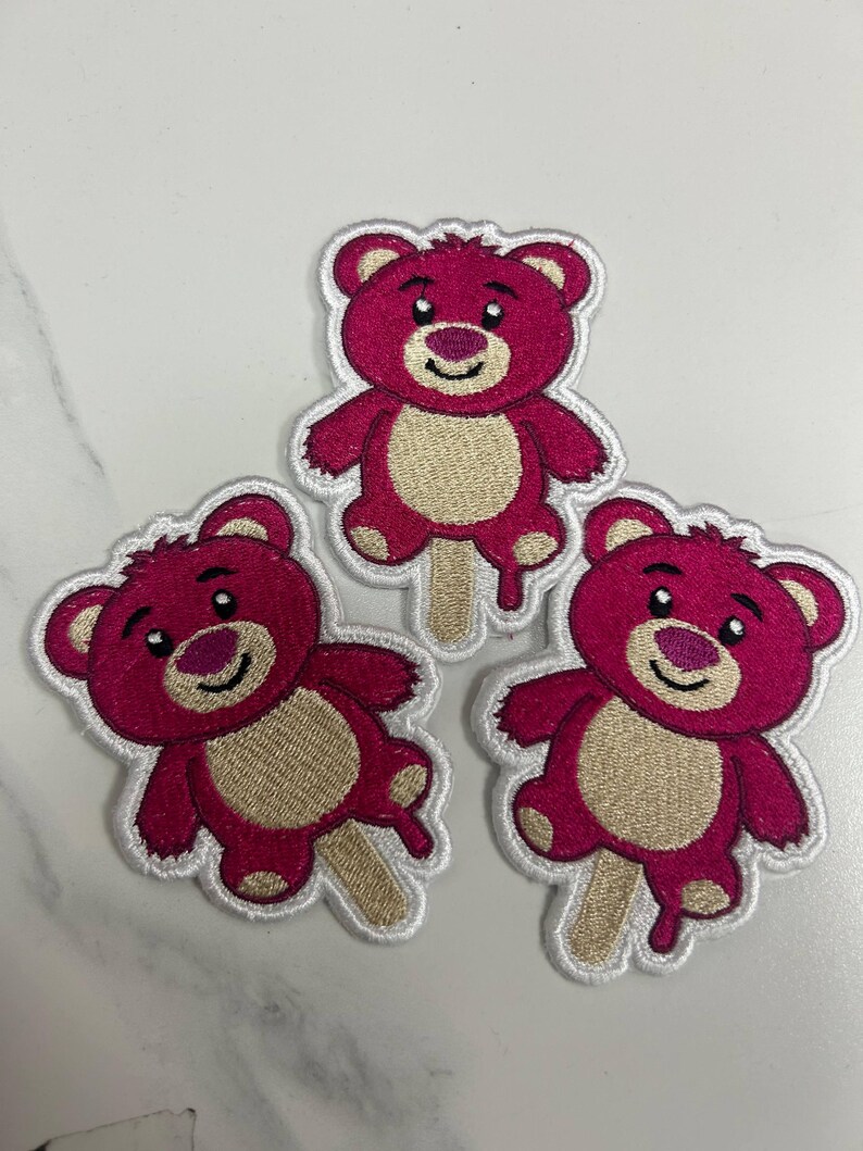 K&ouml;nnte beinhalten: Drei gestickte Aufn&auml;her mit einem rosa Teddyb&auml;r-Design mit beigem Bauch und einem Eisstiel. Der B&auml;r hat ein l&auml;chelndes Gesicht und einen wei&szlig;en Umriss. Die Aufn&auml;her befinden sich auf einer wei&szlig;en Oberfl&auml;che.