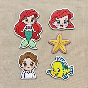 Op de afbeelding: Set van vijf geborduurde patches met personages uit een populaire animatiefilm. Inclusief een zeemeermin met rood haar, een lachend gezicht met rood haar, een gele zeester, een man met bruin haar en een gele vis met blauwe strepen.