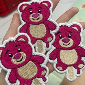 K&ouml;nnte beinhalten: Drei gestickte Aufn&auml;her mit einem rosa Teddyb&auml;r-Design mit hellbraunem Bauch und wei&szlig;er Kontur. Der B&auml;r h&auml;lt einen Eisstiel. Die Aufn&auml;her werden in einer Hand gehalten.