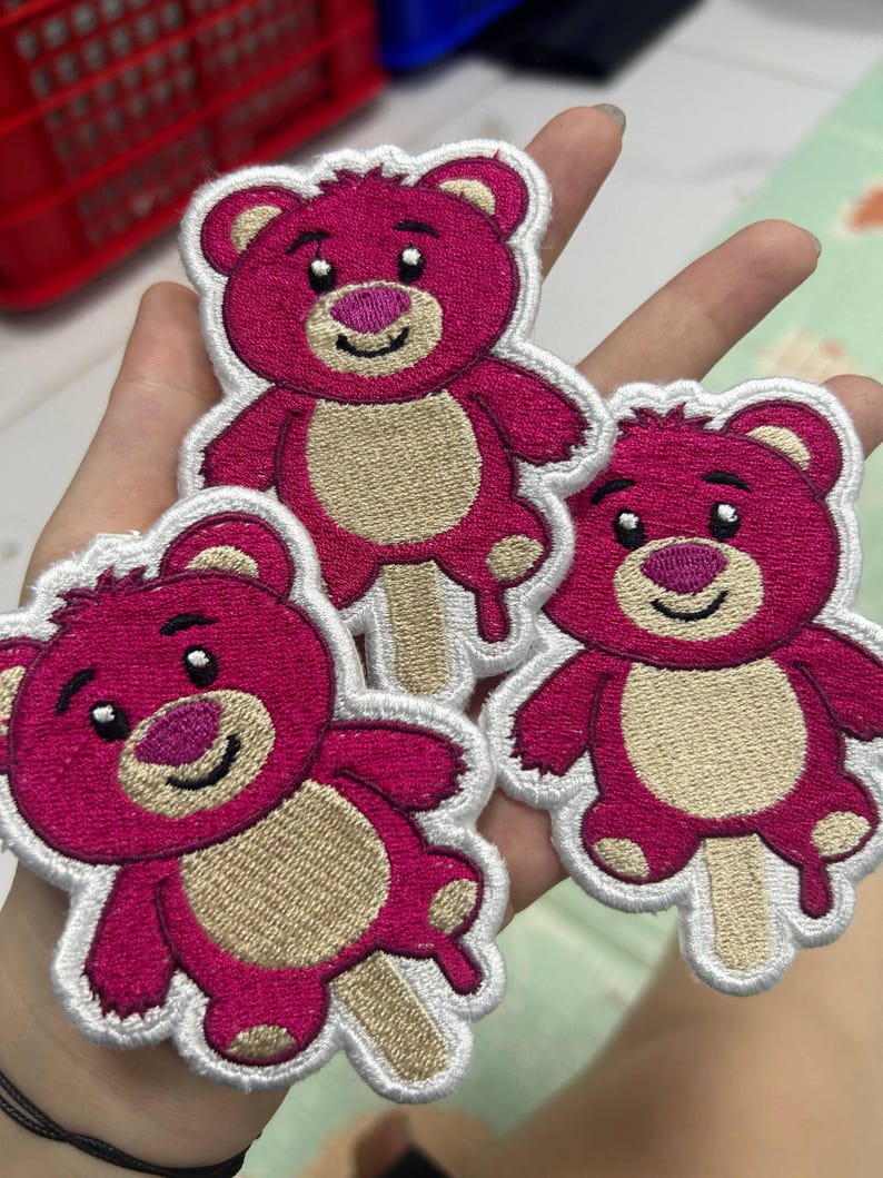 K&ouml;nnte beinhalten: Drei gestickte Aufn&auml;her mit einem rosa Teddyb&auml;r-Design mit hellbraunem Bauch und wei&szlig;er Kontur. Der B&auml;r h&auml;lt einen Eisstiel. Die Aufn&auml;her werden in einer Hand gehalten.