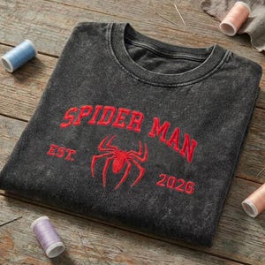 May include: A charcoal gray acid-wash crewneck sweatshirt featuring red embroidered text that reads SPIDER MAN EST. 2026, with a red embroidered spider icon centered below the text. The garment is displayed on a rustic wooden surface.