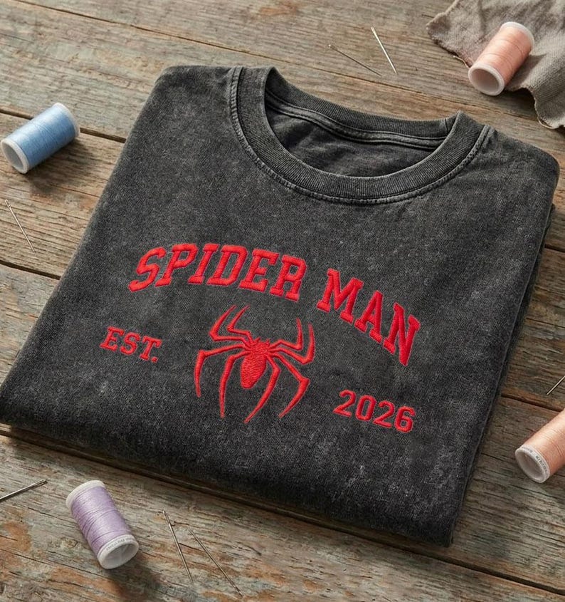 May include: A charcoal gray acid-wash crewneck sweatshirt featuring red embroidered text that reads SPIDER MAN EST. 2026, with a red embroidered spider icon centered below the text. The garment is displayed on a rustic wooden surface.