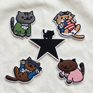 May include: Embroidered patches of cartoon cats in historical attire. The patches depict a grey cat in a green coat, a brown cat with an American flag, a brown cat with a blue headband, and a brown cat in a pink outfit. A black star is in the centre.