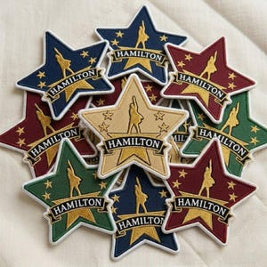 May include: A collection of star-shaped patches in various colors, including navy blue, burgundy, and green. Each patch features the word "HAMILTON" in a banner, a silhouette, and small star accents. The patches are likely for clothing or accessories.