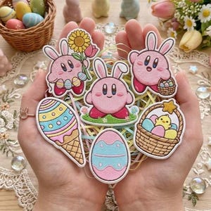 Könnte beinhalten: Eine Sammlung von Aufnähern zum Thema Ostern mit rosa Cartoon-Charakteren mit Hasenohren, Ostereiern und einem Korb mit Eiern. Die Aufnäher sind in verschiedenen Formen und Größen gehalten und weisen farbenfrohe Details und gestickte Ränder auf.