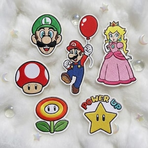 以下が含まれることがあります： スーパーマリオのビデオゲームシリーズのキャラクターとシンボルが刺繍されたパッチ。赤い風船を持ったマリオ、ルイージ、ピーチ姫、赤いキノコ、ファイアフラワー、そして「パワーアップ」の文字が入った黄色の星が含まれています。