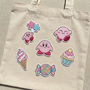 Könnte beinhalten: Eine beigefarbene Tragetasche mit mehreren gestickten Aufnähern. Die Aufnäher zeigen eine rosa Cartoonfigur, ein Eis, einen Cupcake und ein Bonbon mit dem Text "POP STAR". Die Aufnäher sind in verschiedenen Rosa-, Blau- und Gelbtönen gehalten.