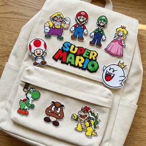 Könnte beinhalten: Beigefarbener Canvas-Rucksack mit bunten Super Mario Charakter-Patches. Zu den Patches gehören Mario, Luigi, Prinzessin Peach und andere. Die Worte "SUPER MARIO" werden in einem mehrfarbigen Patch angezeigt.