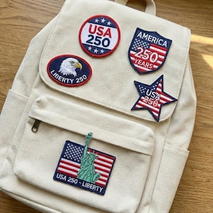 Può includere: Uno zaino color crema decorato con toppe ricamate che celebrano il 250° anniversario degli Stati Uniti. Le toppe includono disegni patriottici, una stella, uno scudo, un'aquila e la Statua della Libertà.