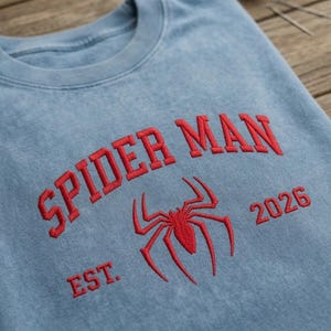 May include: A light blue crewneck sweatshirt featuring red embroidered text that reads SPIDER MAN above a red spider icon, with the text EST. on the left and 2026 on the right of the icon. The fabric has a washed, vintage-style texture.