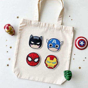 May include: A cream-colored tote bag with four superhero patches: Batman, Captain America, Spider-Man, and Iron Man. Additional superhero-themed accessories include an Iron Man figurine, Captain America shield, and Hulk fist. Gold star confetti is scattered around.