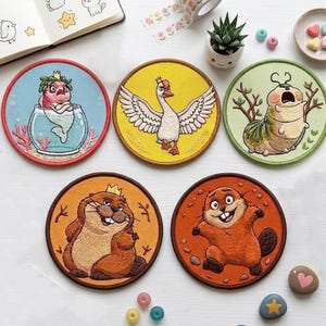 Pode incluir: Uma coleção de cinco patches bordados com desenhos de animais de desenho animado. Os patches apresentam uma sereia, um ganso, uma lagarta, um castor e uma criatura semelhante a um castor. Cada patch é circular com uma borda marrom e fundos coloridos.