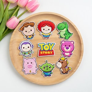 Könnte beinhalten: Eine Sammlung von aufgestickten Aufnähern mit Figuren aus dem Animationsfilm "Toy Story". Die Aufnäher zeigen Woody, Jessie, Rex, Buzz Lightyear, Lotso, Hamm, einen Alien, ein Pferd und das "Toy Story"-Logo. Sie sind auf einem Holztablett angeordnet.