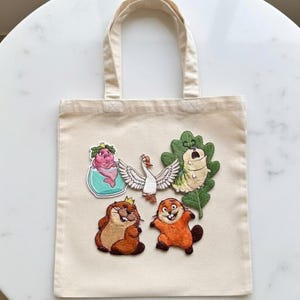 May include: Beige canvas tote bag featuring embroidered cartoon characters. The bag has a pair of brown handles and a rectangular shape. The embroidered characters include a pink creature, a white swan, a green caterpillar, and two brown beavers.