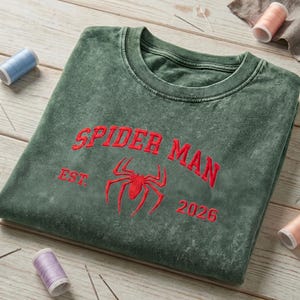 May include: A dark green mineral-washed crewneck sweatshirt featuring red embroidered text that reads SPIDER MAN, EST. 2026, and a central red spider icon. The garment has a ribbed collar and is displayed with sewing thread spools and needles.