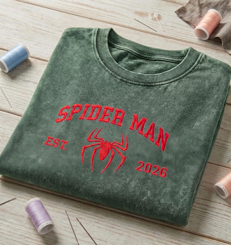 May include: A dark green mineral-washed crewneck sweatshirt featuring red embroidered text that reads SPIDER MAN, EST. 2026, and a central red spider icon. The garment has a ribbed collar and is displayed with sewing thread spools and needles.