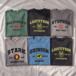 May include: Six crew neck t-shirts in various colors, each featuring a character name, a circular icon, an establishment year, and a location. The shirts display names including Maximoff, Laufeyson, Stephen, Stark, and Odinson with associated graphics.