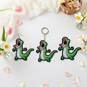 May include: Green cartoon alligator earrings and keychain. The alligator has a white belly, pink tongue, and open mouth. The earrings have silver hooks, and the keychain has a silver ring. The items are on a white surface with flowers.