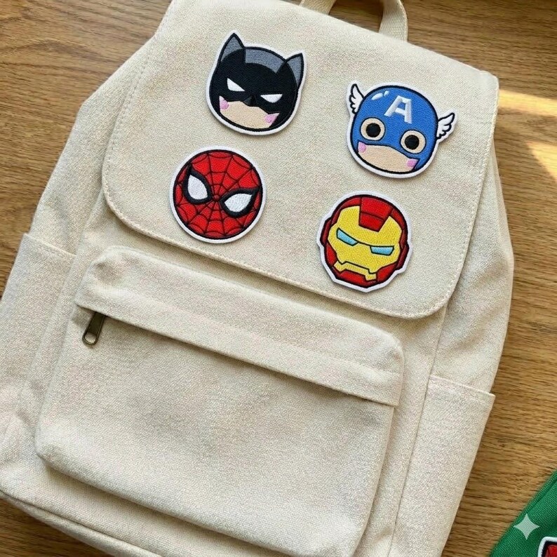 May include: A beige canvas backpack with a front pocket and a top handle. The flap is decorated with four colorful superhero patches: Batman, Captain America, Spider-Man, and Iron Man. The patches are embroidered with black outlines.