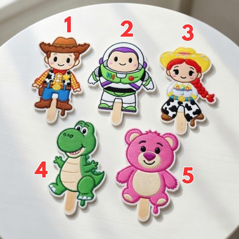 K&ouml;nnte beinhalten: Eine Sammlung von f&uuml;nf Cartoon-Charakter-Patches auf St&auml;ben. Die Charaktere umfassen einen Cowboy, einen Weltraumranger, ein Cowgirl, einen gr&uuml;nen Dinosaurier und einen rosa B&auml;ren. Jeder Patch ist farbenfroh und hat ein detailliertes Design.