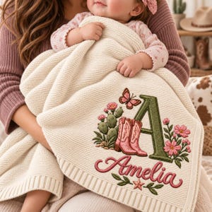 May include: A cream-colored knit baby blanket featuring an embroidered design of a large green letter A, pink cowboy boots, a cactus, a butterfly, and pink flowers. The name Amelia is embroidered in pink script below the letter A.