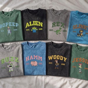 May include: Eight crew neck t-shirts in various colors, each featuring a character from the Toy Story franchise, including Bo Peep, Alien, Rex, Slinky, Buzz, Hamm, Woody, and Jessie, with their names, the text EST. 1995, and a graphic illustration.