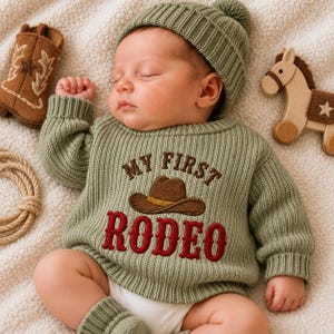 May include: A sage green knitted long-sleeve sweater with a brown cowboy hat graphic and the embroidered text MY FIRST RODEO in red. The sweater is worn by a sleeping infant wearing a matching sage green knit beanie with a pom-pom.