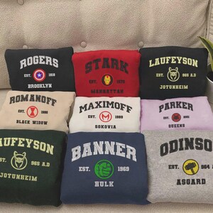 May include: A collection of eight folded sweatshirts in various colors, each featuring a collegiate-style graphic with a character surname, establishment year, and a circular icon representing a superhero, including Rogers, Stark, Laufeyson, Romanoff, Maximoff, Parker, Banner, and Odinson.