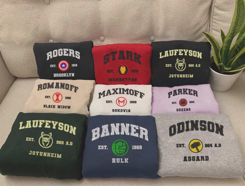 May include: A collection of eight folded sweatshirts in various colors, each featuring a collegiate-style graphic with a character surname, establishment year, and a circular icon representing a superhero, including Rogers, Stark, Laufeyson, Romanoff, Maximoff, Parker, Banner, and Odinson.