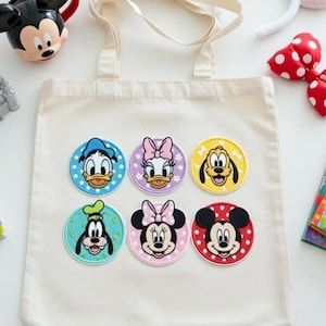 Op de afbeelding: Een crèmekleurige draagtas met zes ronde patches van Disney-personages, waaronder Mickey Mouse, Donald Duck en Goofy. De patches zijn kleurrijk met gedetailleerde gezichten. Andere Disney-accessoires zijn zichtbaar.