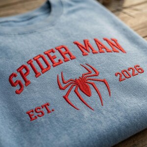 May include: A light blue crewneck sweatshirt featuring red embroidered text that reads SPIDER MAN EST. 2026, centered with a red embroidered spider icon. The fabric has a washed, textured appearance, and the design is stitched directly onto the front.