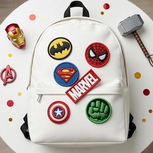 Peut inclure: Un sac à dos blanc orné d'écussons de super-héros, dont Batman, Spider-Man et Captain America. Le mot "MARVEL" est affiché sur un écusson rectangulaire rouge. Des accessoires sur le thème des super-héros sont disposés autour du sac à dos.