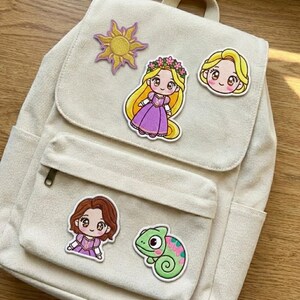 May include: A beige canvas backpack decorated with embroidered patches. Patches include a yellow sun, a princess with long blonde hair and a purple dress, a princess with blonde hair, a princess with brown hair and a green chameleon.