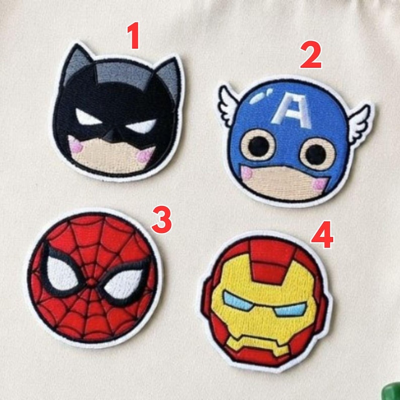 May include: Four embroidered patches featuring superhero faces. Batman is black and gray, Captain America is blue with white wings, Spider-Man is red, and Iron Man is red and gold. Each patch has a white border and is on a light beige background.