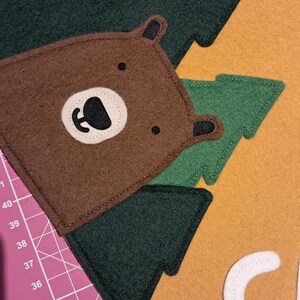 May include: A felt applique featuring a brown bear with a white and black face, green trees, and the letter "C" on an orange background. The applique is likely part of a larger craft project or decoration.