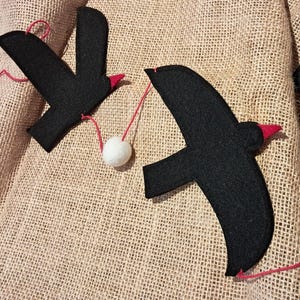 May include: Three black felt bird decorations with red beaks. One bird has a white pom-pom and red string attached. Another has a wooden ring attached. The birds are on a tan burlap surface.
