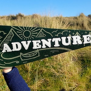 以下が含まれることがあります： 「ADVENTURE」と白く刺繍された文字が書かれた緑色のフェルトのペナント。ペナントには、山、太陽、カヌー、川がスタイリッシュに描かれています。