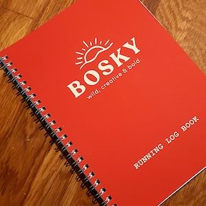 Może przedstawiać: Czerwony notes w spirali z białym słońcem i tekstem "BOSKY wild, creative & bold." oraz "RUNNING LOG BOOK" nadrukowanym na okładce.