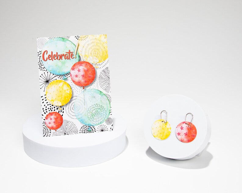 Puede incluir: Un par de pendientes redondos de colores con un dise&ntilde;o de acuarela. Los pendientes est&aacute;n en un expositor blanco. La tarjeta detr&aacute;s de los pendientes dice "Celebrate!" con un dise&ntilde;o de acuarela de globos.