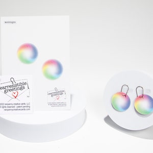 Puede incluir: Un par de pendientes circulares de color arco&iacute;ris sobre un fondo blanco. Los pendientes est&aacute;n hechos de metal y tienen un cierre de gancho. Los pendientes est&aacute;n sobre una superficie blanca. El texto "earresistible greetings" est&aacute; en una tarjeta blanca.