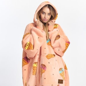 Op de afbeelding: Een oversized hoodie in perzikkleur met een grote zak en een capuchon. De hoodie is bedekt met een patroon van ijshoorntjes met verschillende gekleurde bolletjes ijs.
