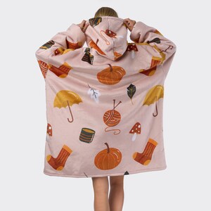 Pu&ograve; includere: Una coperta rosa indossabile con cappuccio. La coperta presenta un motivo ripetitivo di zucche, ombrelli, gomitoli di lana, funghi e calzini.