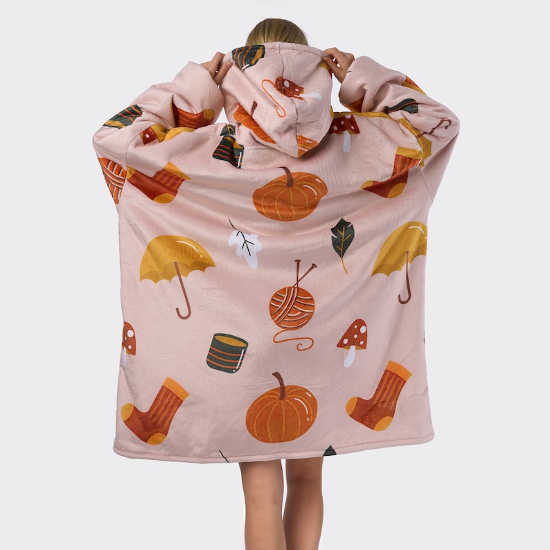 Pu&ograve; includere: Una coperta rosa indossabile con cappuccio. La coperta presenta un motivo ripetitivo di zucche, ombrelli, gomitoli di lana, funghi e calzini.