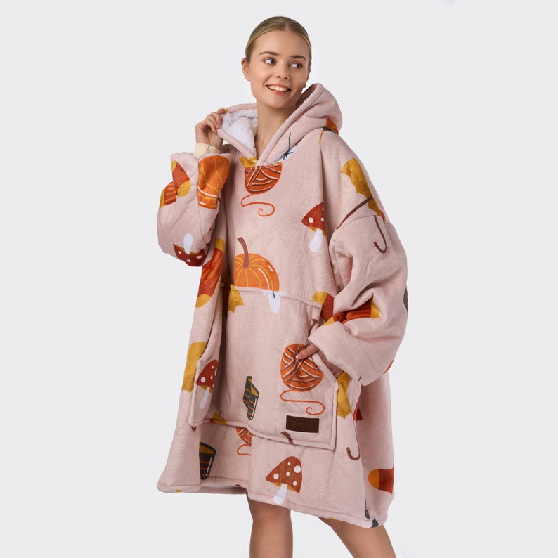Pu&ograve; includere: Una felpa con cappuccio oversize rosa con fodera in sherpa bianca. La felpa presenta un motivo di zucche, funghi e foglie in tonalit&agrave; di arancione, marrone e giallo.