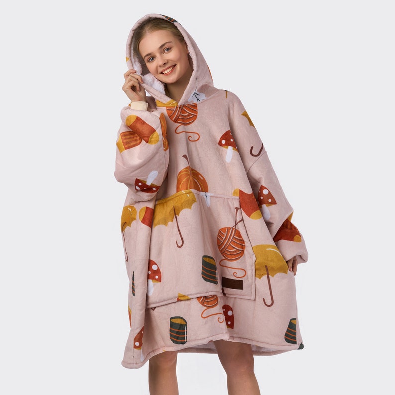 Pu&ograve; includere: Una coperta con cappuccio rosa con un motivo di funghi, zucche, gomitoli di lana e ombrelli. La coperta ha grandi tasche sul davanti.