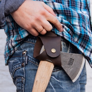 May include: A person wearing a blue plaid shirt and blue jeans is carrying a small axe with a brown leather sheath. The axe has a wooden handle and a gray metal head.