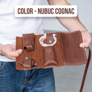 Könnte beinhalten: Ein brauner Reiseorganizer aus Leder mit mehreren Taschen und Fächern zum Verstauen von Kleinigkeiten wie Kabeln, Ladegeräten und einem Telefon. Der Organizer ist geöffnet und flach ausgelegt, so dass das Innere sichtbar ist. Der Text "COLOR - NUBUC COGNAC" ist oben im Bild zu sehen.